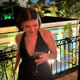 Woman in black dress holding a drink on a balcony overlooking a pool at night with illuminated tropical surroundings