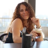 Woman with curly reddish-brown hair smiling at a table with a beverage, city skyline visible through window behind her