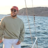 Man wearing red cap and sunglasses standing on a sailboat near a rocky coastline