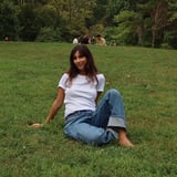 Woman in white t-shirt and jeans sitting on grass in a park with trees and people in the background